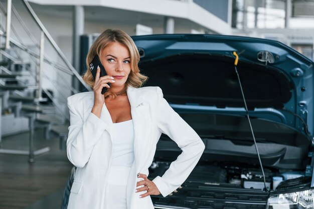 Photo talking by phone woman in formal clothes is indoors in the autosalon