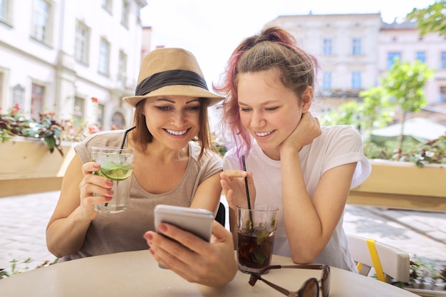 Talking and drinking soft drinks mother daughter teenager, smiling women sitting in outdoor cafe, looking at smartphone. Friendship and communication of parent and child of teenager