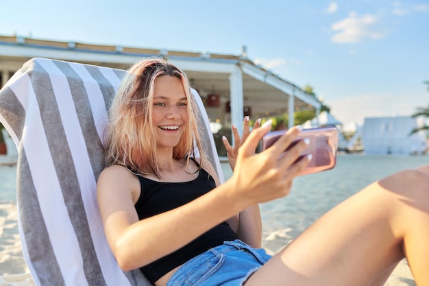 Talking female teenager in beach sun lounger making video call using smartphone. Vacation, weekend at sea, travel tourism technology concept