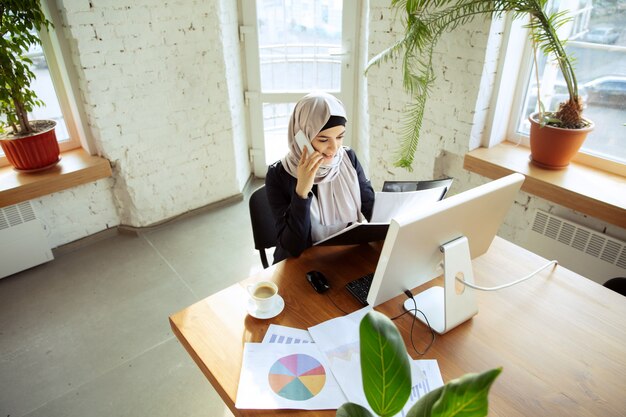 Parlare al telefono durante la revisione dei documenti. bella donna d'affari araba che indossa l'hijab mentre lavora in openspace o in ufficio. concetto di occupazione, libertà nel settore degli affari, successo, soluzione moderna.