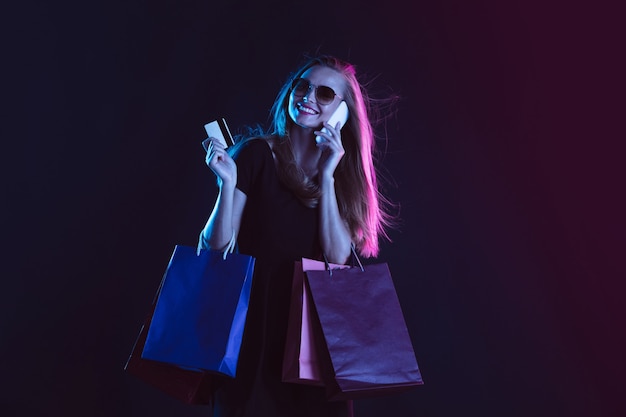ショッピングバッグとカードで話す電話。暗い背景にネオンの光の若い女性の肖像画。人間の感情、ブラックフライデー、サイバーマンデー、購入、販売、財務の概念。