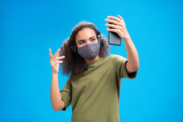 Talking on video call young African American girl using her smartphone with headphones on in olive t-shirt, reusable face mask