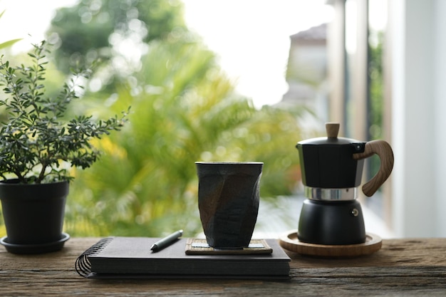 Tall black coffee mug and moka pot and notebook diary on old brown wooden table