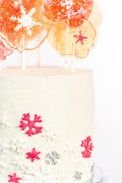 Tall round cake with Italian buttercream frosting decorated with fondant snowflakes and topped with large pink and white lollipops on a white background.