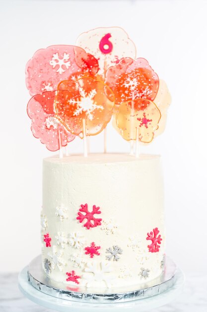 Tall round cake with Italian buttercream frosting decorated with fondant snowflakes and topped with large pink and white lollipops on a white background.