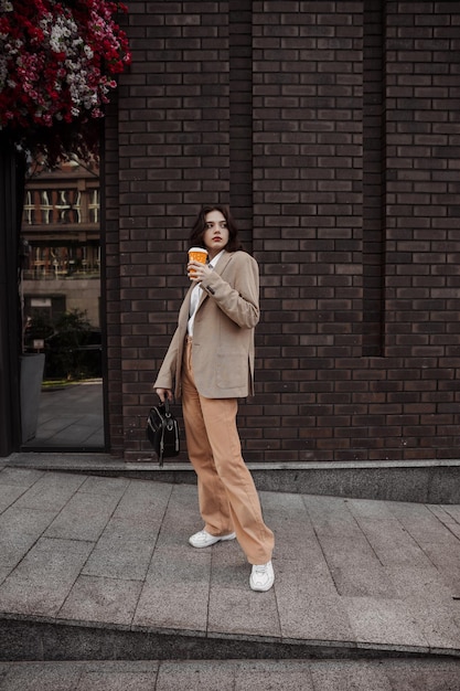 Ragazza alta ed elegante con caffè in una giacca oversize beige e pantaloni arancioni sullo sfondo del muro di mattoni