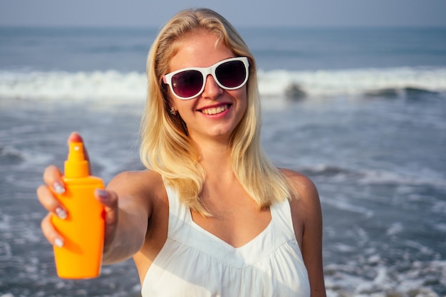 Tan woman applying sun protection lotion in white dress and glasses on the beach