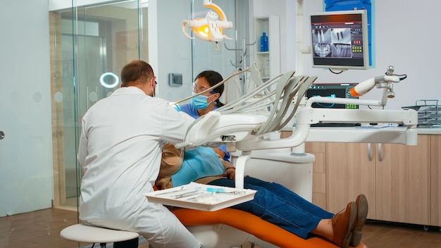 Tandarts die tanden behandelt aan hogere vrouwenpatiënt in kliniek die op stoel met open mond ligt. Arts en verpleegster werken samen in een modern stomatologisch kantoor met een beschermingsmasker