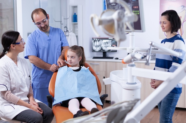 Tandarts in gesprek met ouder over mondhygine van kinderen voor gezondheid in tandartspraktijk. Kind met haar moeder tijdens tandencontrole met stomatolog zittend op een stoel.