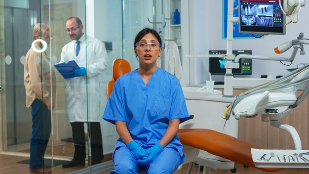 Tandartsassistente die naar de camera kijkt en met patiënten spreekt over mondhygiëne. Stomatoloog praten op webcam zittend op een stoel in stomatologische kliniek met arts op de achtergrond die op de computer werkt