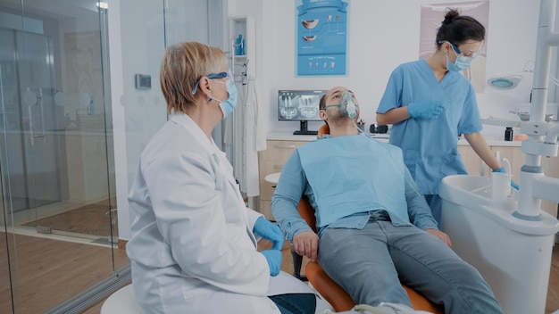 Tandheelkundige assistent die zuurstofmasker op het gezicht van de patiënt zet om anesthesie te doen vóór de chirurgische ingreep. Mondzorgspecialist die verdoving geeft aan man met kiespijn en boorgereedschap gebruikt.