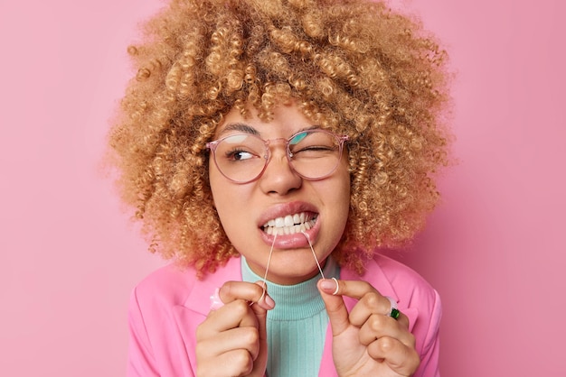 Foto tandheelkundige zorgconcept mooie jonge vrouw met krullend borstelig haar reinigt tanden met tandzijde draagt transparante bril geïsoleerd over roze achtergrond laat zien hoe correct flossen