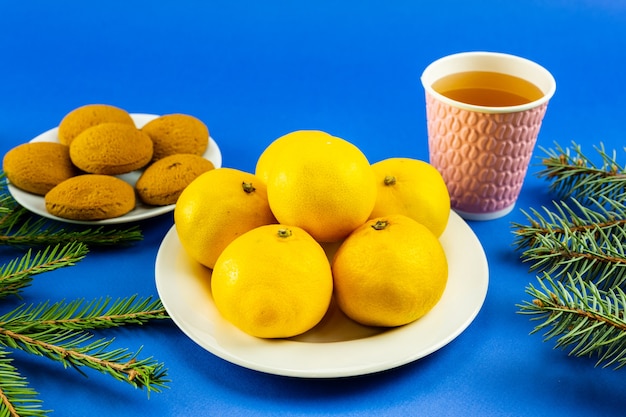 Tangerines on a white plate, fir branches, a pink glass and cookies on a blue background.