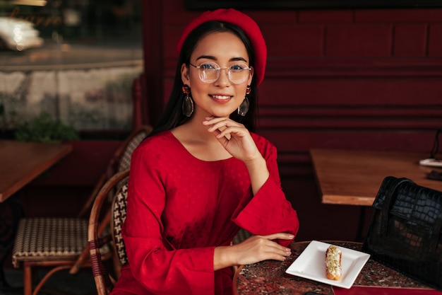 Tanned Asian woman in red beret, bright dress and eyeglasses smiles, sits in gorgeous cafe and looks into camera