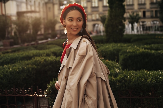 Una bella donna bruna abbronzata con un berretto rosso alla moda e un trench beige alla moda sorride, si guarda indietro e cammina nel parco