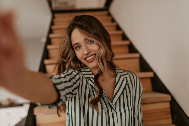 Tanned blueeyed woman in striped silk green and white shirt smiles sincerely and takes selfie at h