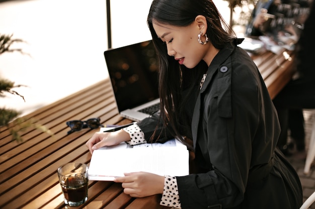 Tanned brunette Asian woman in black trench coat smiles, sits at wooden desk with glass of coffee and reads book outside