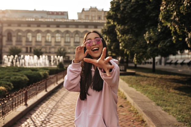 Tanned brunette Asian woman in pink hoodie and stylish sunglasses reaches to camera and smiles widely outside