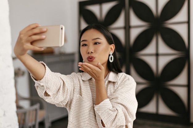 Tanned brunette woman in beige jacket takes selfie in cozy room Pretty lady in good mood holds phone and blows kiss