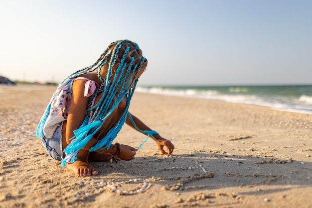 Tanned enthusiastic slim girl with bright african braids in summer pink summer suit plays on beach with seashells near sea with foamy fluffy waves under warm sunny golden sunset