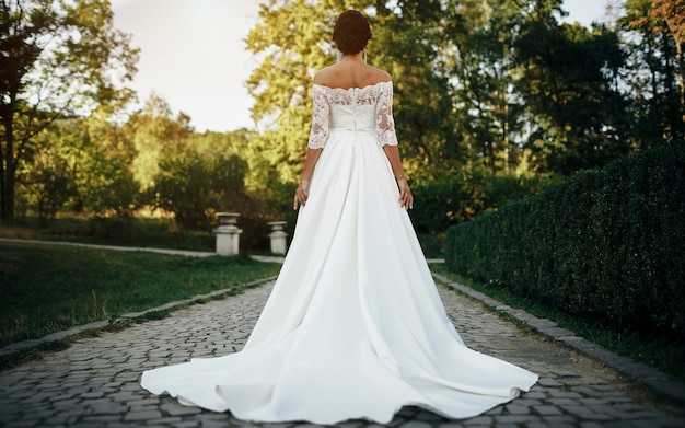 A tanned girl bride in a chic white wedding dress with bare shoulders stands with her back turned in the park on the alley