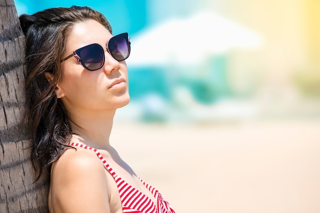 Tanned girl in sunglasses by palm tree. The girl takes sun baths. Recreation and tourism concept.