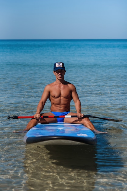 L'uomo abbronzato si siede sulla spiaggia vicino alla sua tavola da surf e guarda alla telecamera.