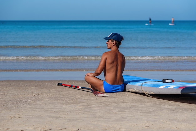 Tanned man sits on the beach near his surfboard and looks to the sea. 