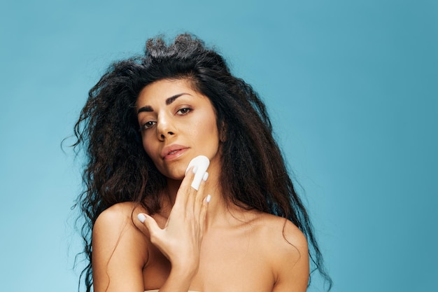 Tanned sexy beautiful curly latin lady holding cotton pad for\
face cleansing face with toner after shower over blue studio\
background pensive woman enjoying fresh moisturized face skin copy\
space