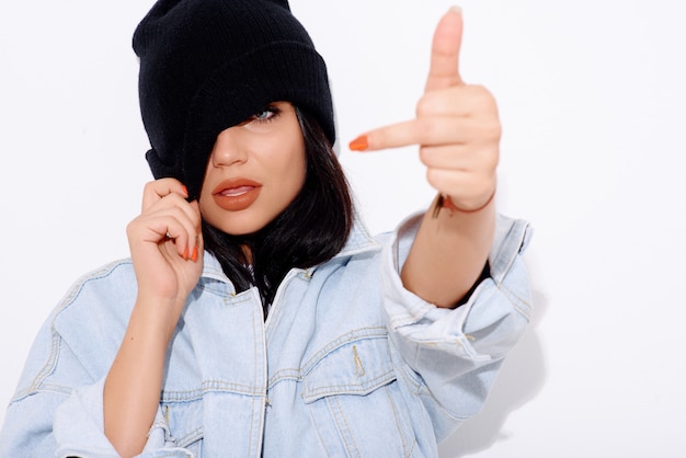 Tanned woman in a denim jacket and shorts with a hat on a white wall shows  finger