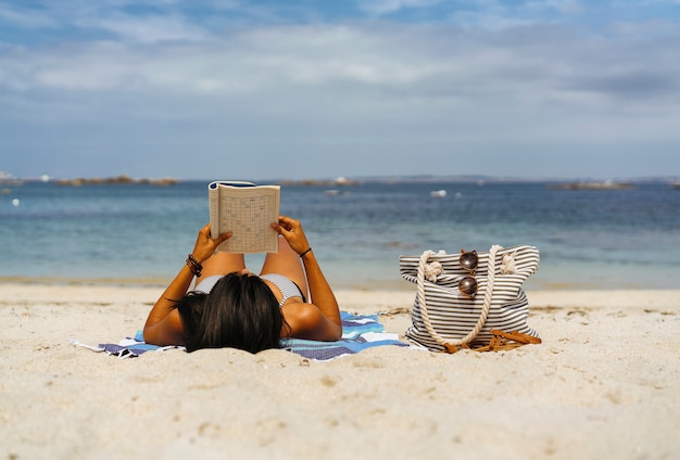 Foto donna abbronzata leggendo e godendo sulla spiaggia dell'oceano