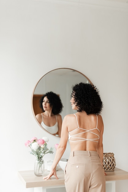 Photo tanned woman with a bare back and black curly hair stands in front of a mirror