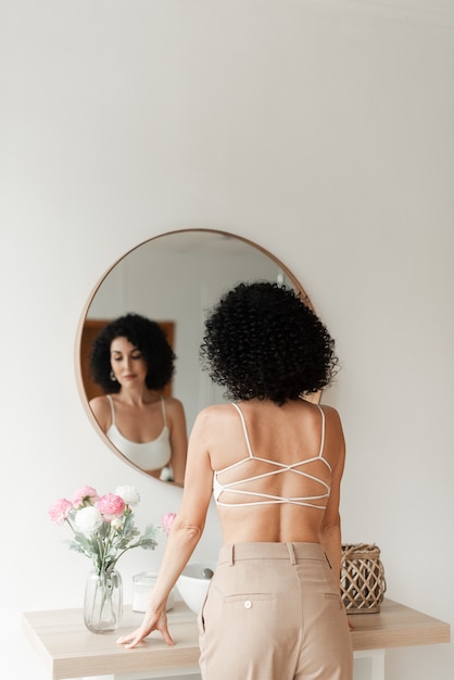 Photo tanned woman with a bare back and black curly hair stands in front of a round mirror