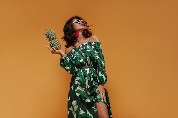 Tanned wonderful girl with wavy brunette hairstyle and red earrings in sunglasses and green dress holding pineapple and posing