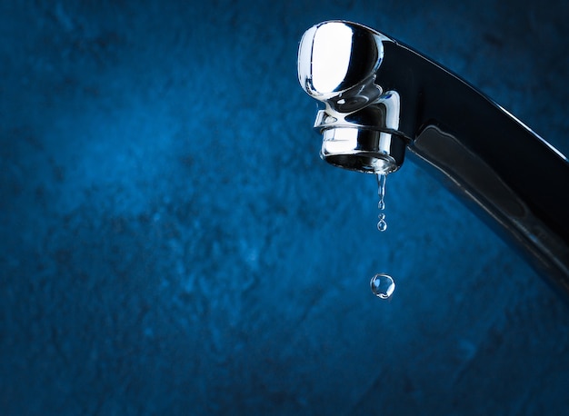 Tap with dripping water close-up on blue background. Water consumption concept