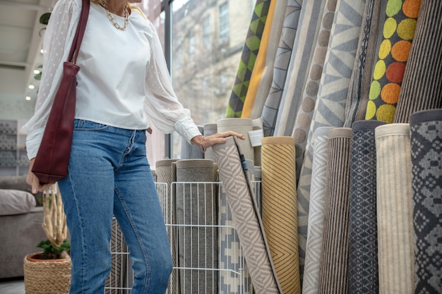 Tapijten, assortiment. Vrouw in een witte blouse en spijkerbroek die op de tapijtafdeling van een meubelwinkel staat en een kleed met haar hand vasthoudt, haar gezicht is niet zichtbaar.