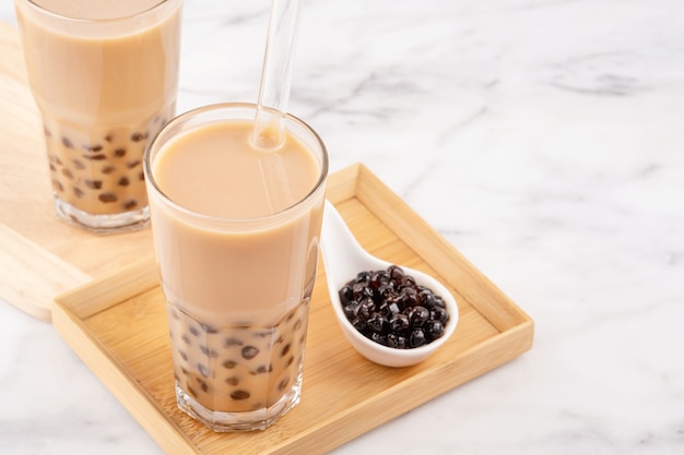 Tapioca pearl ball bubble milk tea, popular Taiwan drink, in drinking glass with straw on marble white table and wooden tray