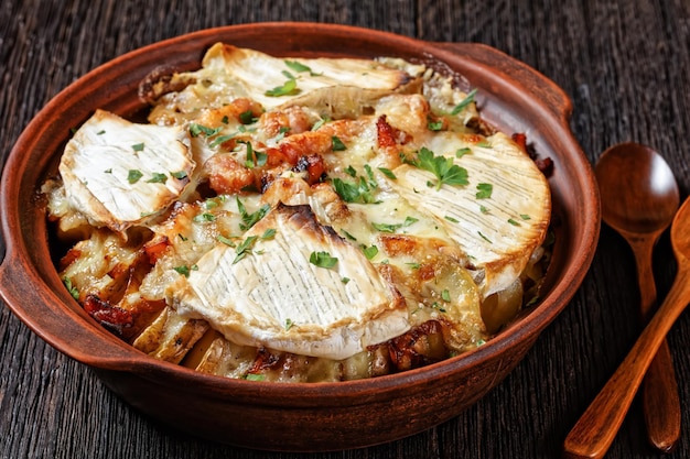 Tartiflette, French Potato, Bacon, and Cheese Casserole in a clay dish  on a dark wooden table with spoons,  french cuisine