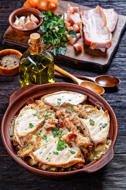 Tartiflette, French Potato, Bacon, and Cheese Casserole in a clay dish  on a dark wooden table with spoons, vertical view, french cuisine