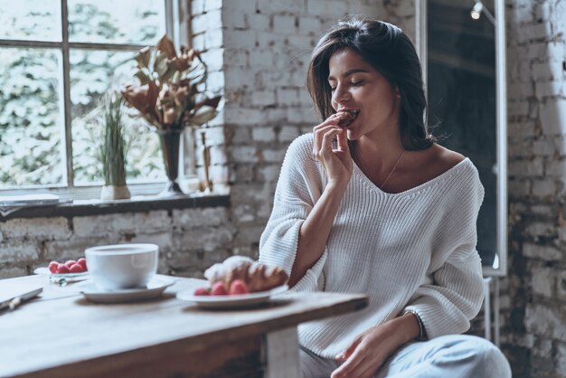 Tastes delicious! Attractive young woman keeping eyes closed and eating croissant 