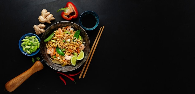 Вкусная аппетитная азиатская лапша с овощами и креветками на сковороде на темной поверхности