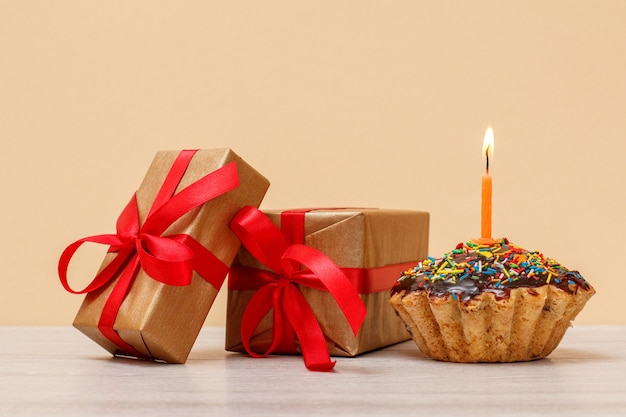 Tasty birthday muffin with chocolate glaze and caramel, decorated with burning festive candle and gift boxes on beige background. Happy birthday minimal concept.