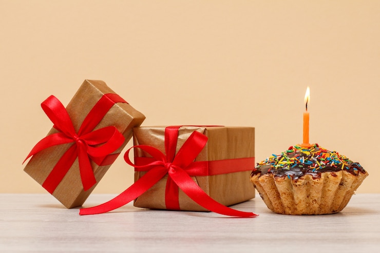 Photo tasty birthday muffin with chocolate glaze and caramel, decorated with burning festive candle and gift boxes on beige background. happy birthday minimal concept.