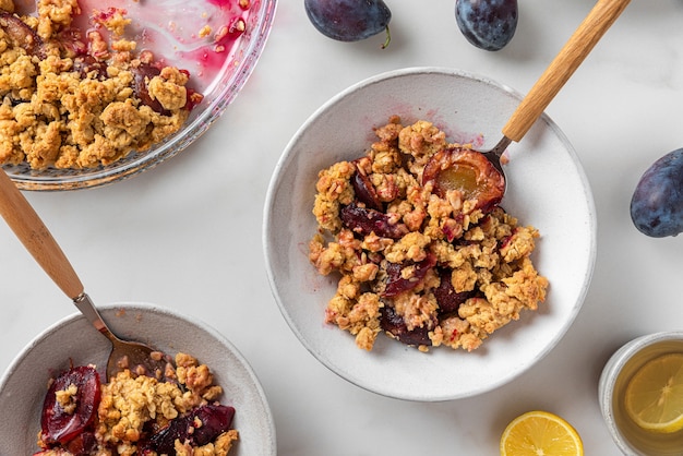 Tasty breakfast. Plum crumble dessert in plates with spoons and tea cup with lemon on white background. Top view