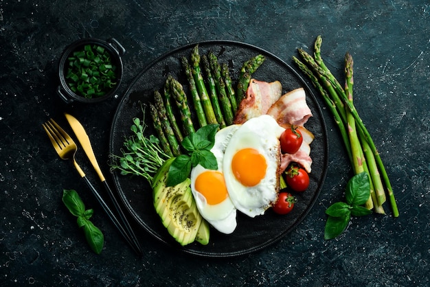 Tasty breakfast Sunny side up eggs asparagus bacon and avocado On a black stone plate