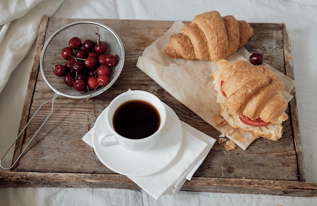 Gustosa colazione con croissant freschi, caffè, ciliegie su un vassoio di legno. croissant abbondante con pomodoro e formaggio. espresso sul vassoio della colazione