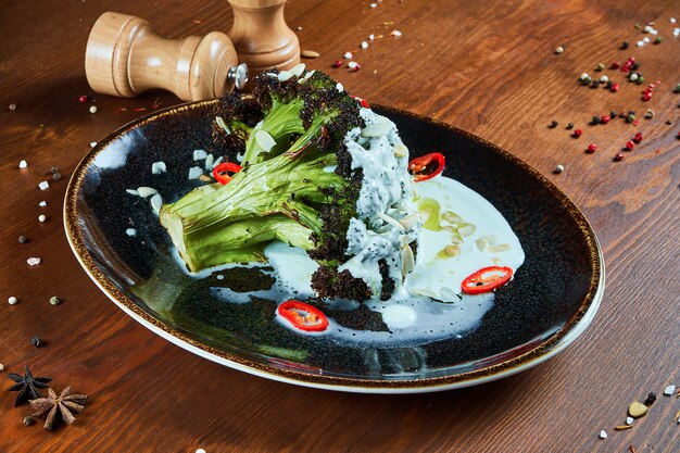 Фото Вкусная брокколи с йогуртовым соусом, острым перцем и специями на стильной черной тарелке. ресторан, где подают еду. вегетарианская еда .. деревянный стол.