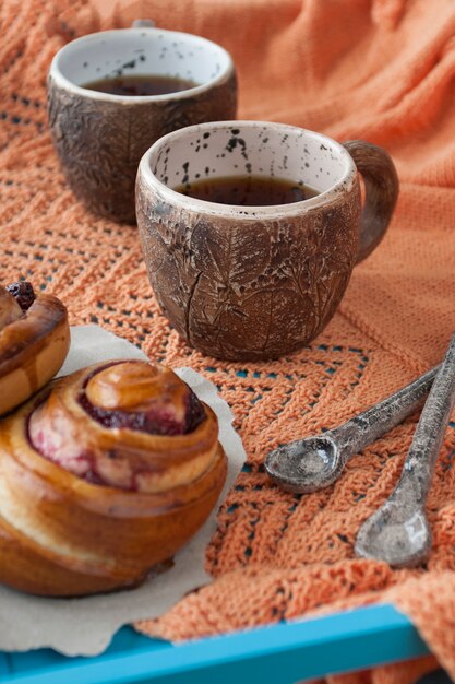 Tasty buns with jam and two cups of tea on blue wood. Next there are two spoons and a bright orange knitted plaid.