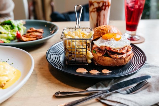 Tasty burger with fried egg served with fries in black plate on wooden table.