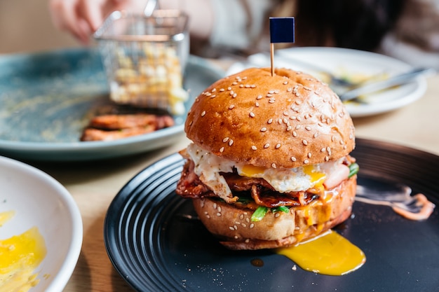 Tasty burger with fried egg and yolk served with fries in black plate on wooden table.